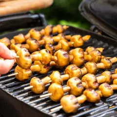 Leckere Champignons grillen auf dem Rost. Eine Hand steckt einen weiteren Spieß mit Pilzen auf den Grill.