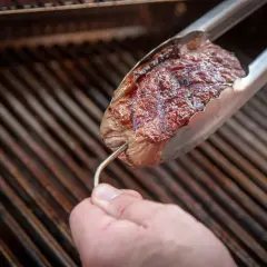 Ein Steak wird mit einer Zange über einem Grill gehalten. Ein Thermometer wird eingeführt, um die Kerntemperatur Rind zu prüfen.