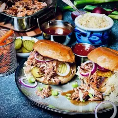 Zwei saftige Pulled Pork Burger, zubereitet im Sous Vide Verfahren, mit Krautsalat, Gurken und roten Zwiebeln. Dazu Pommes und BBQ-Sauce.