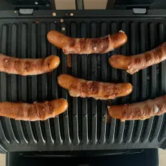 Sechs **Berner Würste** brutzeln auf einem Kontaktgrill. Die Würste sind mit Speck umwickelt und werden goldbraun.