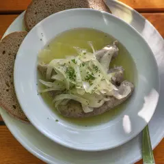Drei **blaue Zipfel** in einer Schüssel mit Zwiebeln und Brühe, dazu zwei Scheiben dunkles Brot auf einem Holztisch.