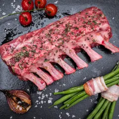 Rohes Lammkarree mit Kräutern und Gewürzen, bereit zum Lammkarree grillen. Dazu grüne Bohnen im Speckmantel, Kirschtomaten und gegrillte Zwiebel.