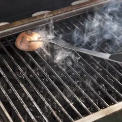 Eine Zwiebel wird mit einer Zange auf dem Grillrost platziert, um den Grill ausbrennen zu lassen. Rauch steigt auf.
