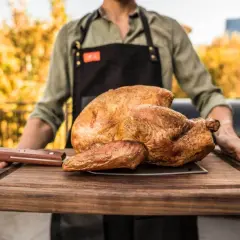 Ein Mann mit Schürze trägt ein perfekt gebratenes Huhn auf einem Holzbrett. Die Frage "Huhn kochen wie lange?" beschäftigt viele Hobbyköche.
