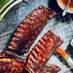 Saftige spareribs 321, glasiert mit BBQ-Sauce, liegen auf einem Grillrost. Frühlingszwiebeln und eine Sauce-Schale daneben.