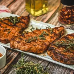 Saftige Nackensteaks vom Grill, garniert mit frischem Thymian. Daneben eine Schale Gewürzmischung und ein Glas Chiliflocken.