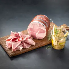 Ein runder Bierschinken liegt auf einem Holzbrett, daneben aufgeschnittene Scheiben und ein Glas mit eingelegtem Gemüse.