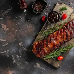 Saftige Rippchen, perfekt mariniert und gegrillt, liegen auf einem Holzbrett. Dazu gibt es BBQ-Sauce, Rosmarin und Tomaten.