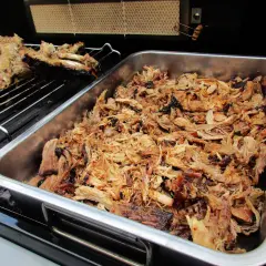 Saftiges pulled pork liegt in einer Metallschale auf dem Gasgrill. Daneben auf dem Rost ein Teil eines Rippchenbratens.