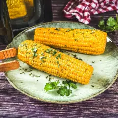 Zwei goldgelbe Maiskolben, frisch aus dem Airfryer, liegen auf einem Teller, garniert mit Kräutern. Perfekt für einen leckeren Snack!