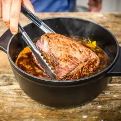 Ein saftiges Stück Fleisch wird mit einer Zange aus einem Topf gehoben, in dem es in einer dunklen Soße schmort. Das langsame Garen, das Schmoren, macht es zart.