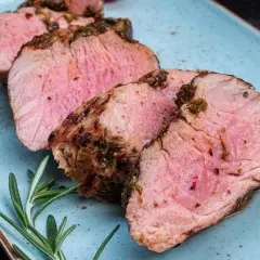 Saftiger schweinelummer, perfekt gegrillt und mit Kräutern verfeinert, liegt auf einem blauen Teller. Ein Rosmarinzweig und eine halbe Tomate garnieren das Gericht.