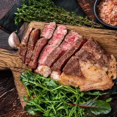 Perfekt gegartes Côte de Boeuf sous vide, in Scheiben geschnitten, mit frischem Grünzeug und Rosmarinzweig auf Holzbrett.