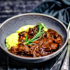 Ein Teller mit cremigem Kartoffelpüree und einem reichhaltigen Stracotto di manzo, garniert mit Rosmarin. Daneben liegen silberne Gabel und Löffel.