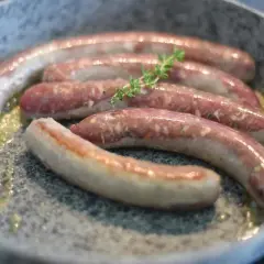 Fünf rohe Bratwürste brutzeln in einer Pfanne mit Butter. Ein Zweig Thymian liegt auf den Würsten, ein Hinweis auf ein köstliches wildbratwurst rezept.