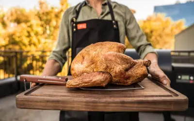 Ein Mann mit Schürze trägt ein perfekt gebratenes Huhn auf einem Holzbrett. Die Frage "Huhn kochen wie lange?" beschäftigt viele Hobbyköche.