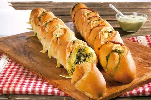 Zwei gefüllte Baguettes zum Grillen, eines mit Kräutern und Käse, das andere mit Pesto und Tomaten, daneben eine Schüssel Dip.