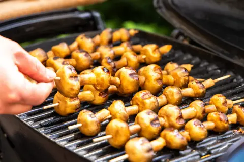 Leckere Champignons grillen auf dem Rost. Eine Hand steckt einen weiteren Spieß mit Pilzen auf den Grill.
