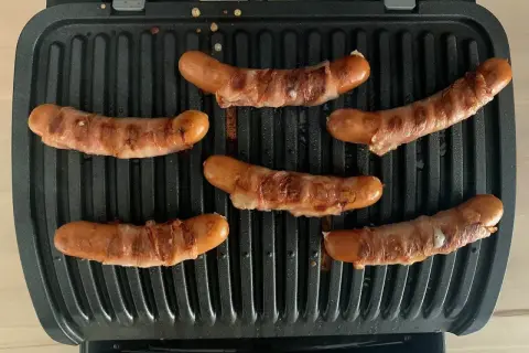 Sechs **Berner Würste** brutzeln auf einem Kontaktgrill. Die Würste sind mit Speck umwickelt und werden goldbraun.
