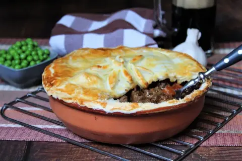 Herzhaftes Steak Pie mit goldbrauner Kruste, dane eine Schüssel Erbsen und ein Glas dunkles Bier.