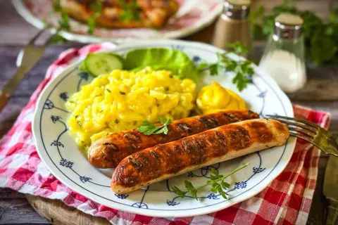 Zwei gebratene Bratwürste mit Kartoffelsalat, Gurkenscheiben, Salatblatt und Senf. Die Bratwurst Zutaten sind hier perfekt kombiniert.