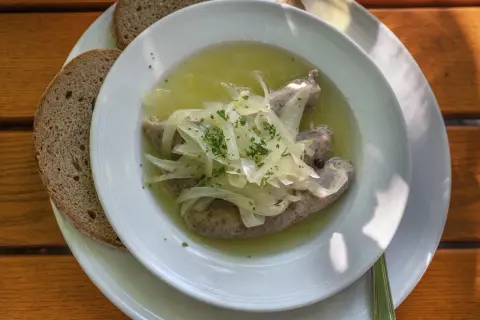 Drei **blaue Zipfel** in einer Schüssel mit Zwiebeln und Brühe, dazu zwei Scheiben dunkles Brot auf einem Holztisch.