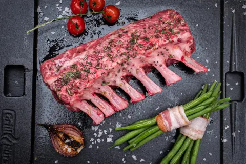 Rohes Lammkarree mit Kräutern und Gewürzen, bereit zum Lammkarree grillen. Dazu grüne Bohnen im Speckmantel, Kirschtomaten und gegrillte Zwiebel.