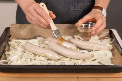 Bratwurst im Backofen: Mit einem Pinsel wird die Wurst mit Öl bestrichen, bevor sie auf Zwiebeln im Ofen gart.