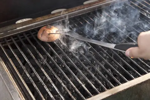 Eine Zwiebel wird mit einer Zange auf dem Grillrost platziert, um den Grill ausbrennen zu lassen. Rauch steigt auf.