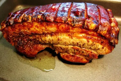 Perfekt gegrillter Schweinebauch mit knuspriger Schwarte liegt in einer Bratpfanne. Der Bratensaft glänzt daneben.
