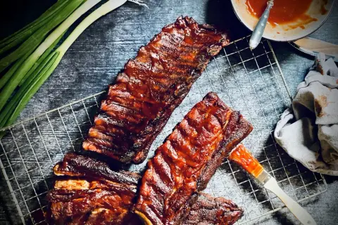 Saftige spareribs 321, glasiert mit BBQ-Sauce, liegen auf einem Grillrost. Frühlingszwiebeln und eine Sauce-Schale daneben.