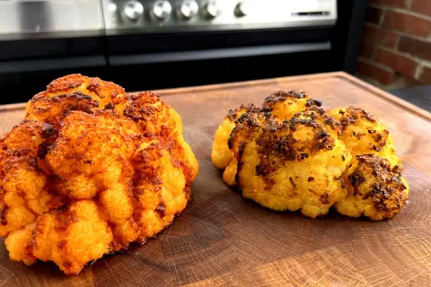 Zwei perfekt gegrillte Blumenkohlröschen liegen auf einem Holzbrett. Sie sind goldbraun und leicht angekohlt, was auf köstliches blumenkohl grillen hindeutet.