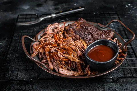 Saftiges pulled pork mit BBQ-Sauce und Beilagen auf einem Teller. Ein Gabel liegt daneben.