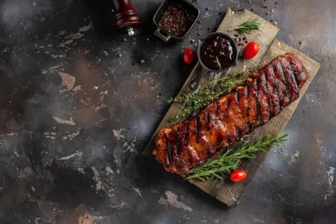 Saftige Rippchen, perfekt mariniert und gegrillt, liegen auf einem Holzbrett. Dazu gibt es BBQ-Sauce, Rosmarin und Tomaten.