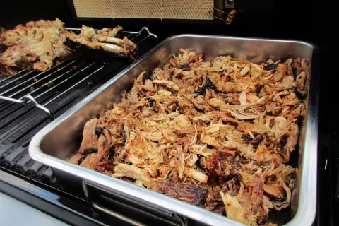 Saftiges pulled pork liegt in einer Metallschale auf dem Gasgrill. Daneben auf dem Rost ein Teil eines Rippchenbratens.