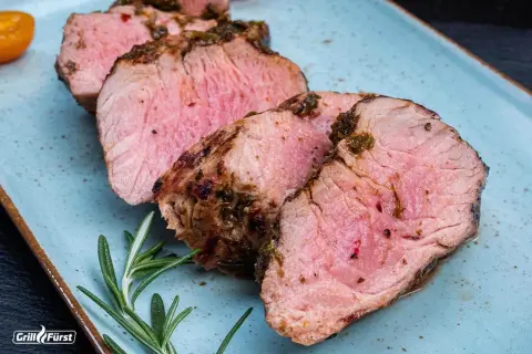 Saftiger schweinelummer, perfekt gegrillt und mit Kräutern verfeinert, liegt auf einem blauen Teller. Ein Rosmarinzweig und eine halbe Tomate garnieren das Gericht.