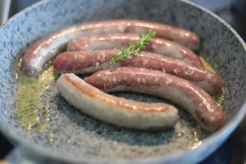 Fünf rohe Bratwürste brutzeln in einer Pfanne mit Butter. Ein Zweig Thymian liegt auf den Würsten, ein Hinweis auf ein köstliches wildbratwurst rezept.