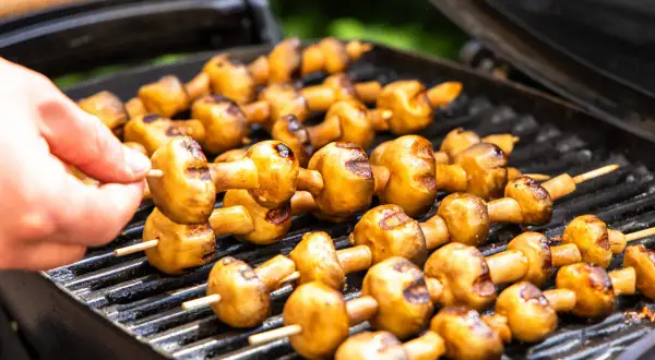 Leckere Champignons grillen auf dem Rost. Eine Hand steckt einen weiteren Spieß mit Pilzen auf den Grill.