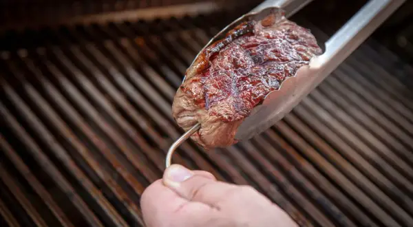 Ein Steak wird mit einer Zange über einem Grill gehalten. Ein Thermometer wird eingeführt, um die Kerntemperatur Rind zu prüfen.