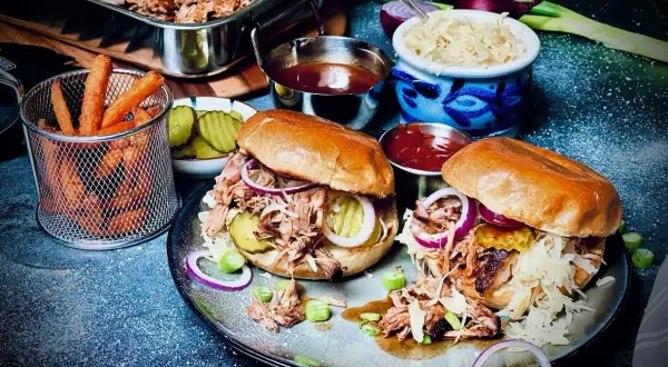 Zwei saftige Pulled Pork Burger, zubereitet im Sous Vide Verfahren, mit Krautsalat, Gurken und roten Zwiebeln. Dazu Pommes und BBQ-Sauce.