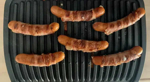 Sechs **Berner Würste** brutzeln auf einem Kontaktgrill. Die Würste sind mit Speck umwickelt und werden goldbraun.