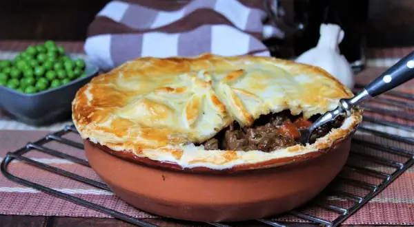 Herzhaftes Steak Pie mit goldbrauner Kruste, dane eine Schüssel Erbsen und ein Glas dunkles Bier.