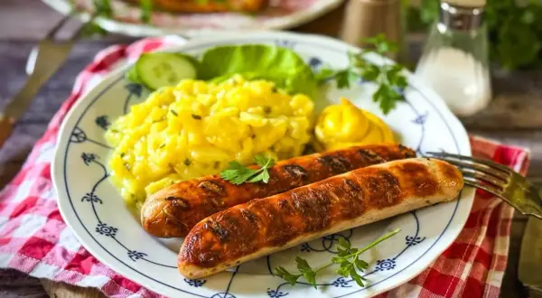 Zwei gebratene Bratwürste mit Kartoffelsalat, Gurkenscheiben, Salatblatt und Senf. Die Bratwurst Zutaten sind hier perfekt kombiniert.