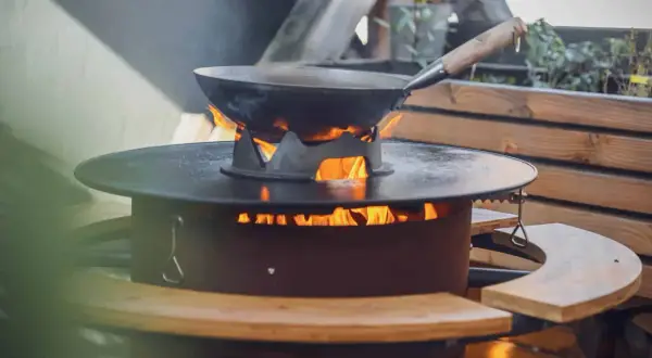 Ein Wok auf einem Feuerring, der an eine Plancha erinnert, wird draußen erhitzt.