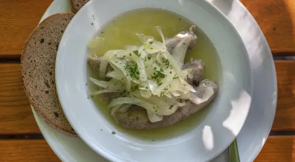 Drei **blaue Zipfel** in einer Schüssel mit Zwiebeln und Brühe, dazu zwei Scheiben dunkles Brot auf einem Holztisch.