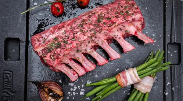 Rohes Lammkarree mit Kräutern und Gewürzen, bereit zum Lammkarree grillen. Dazu grüne Bohnen im Speckmantel, Kirschtomaten und gegrillte Zwiebel.
