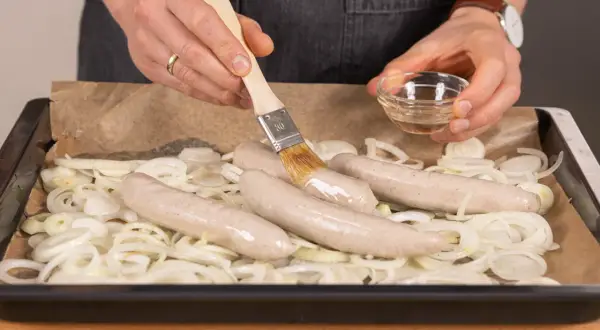 Bratwurst im Backofen: Mit einem Pinsel wird die Wurst mit Öl bestrichen, bevor sie auf Zwiebeln im Ofen gart.