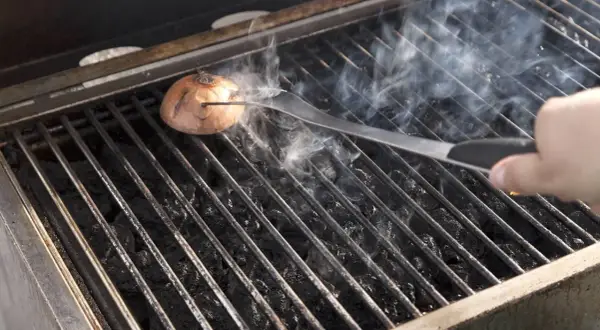 Eine Zwiebel wird mit einer Zange auf dem Grillrost platziert, um den Grill ausbrennen zu lassen. Rauch steigt auf.