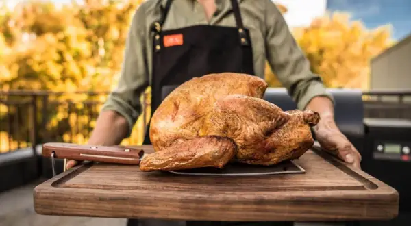 Ein Mann mit Schürze trägt ein perfekt gebratenes Huhn auf einem Holzbrett. Die Frage "Huhn kochen wie lange?" beschäftigt viele Hobbyköche.
