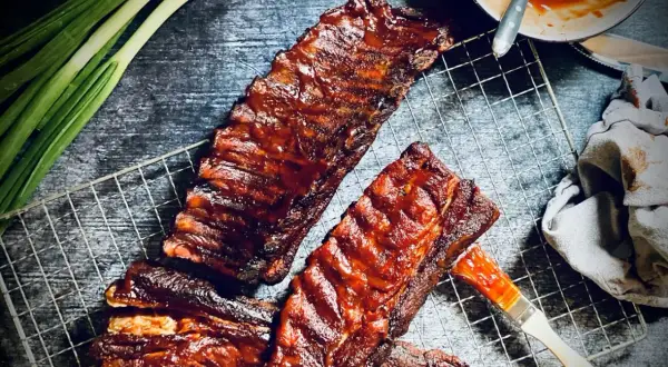 Saftige spareribs 321, glasiert mit BBQ-Sauce, liegen auf einem Grillrost. Frühlingszwiebeln und eine Sauce-Schale daneben.